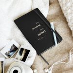 Schwarzes Notizbuch mit heller Typografie und Nudelholzmotiv, schräg auf hellem Stoff fotografiert