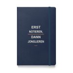 Marineblaues Hardcover-Notizbuch mit cremefarbener Typografie „Erst notieren, dann jonglieren“ und orangefarbenen Akzenten ohne Hintergrund.