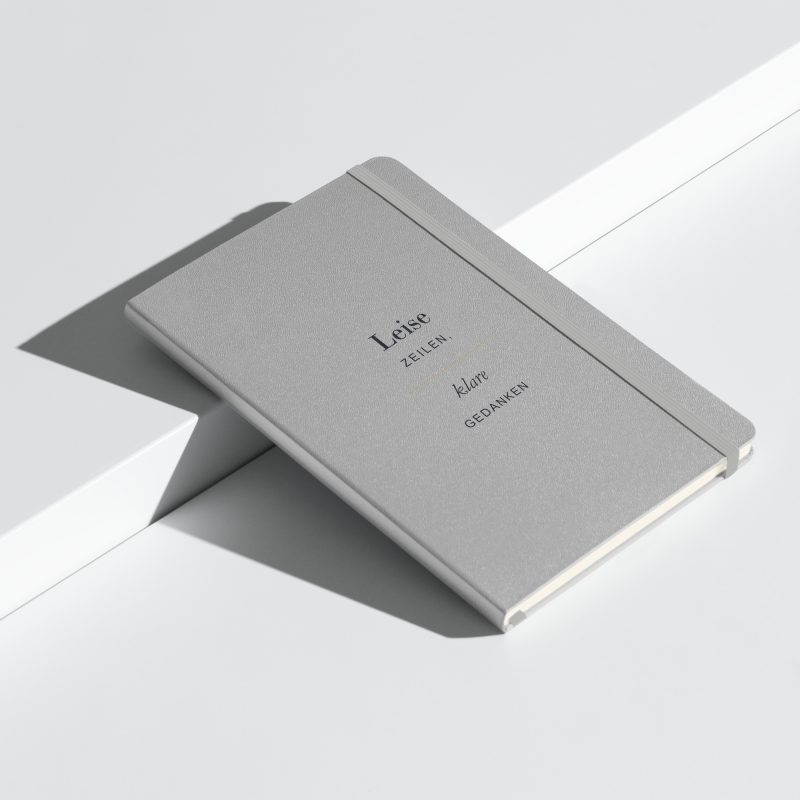 Silberfarbenes Hardcover-Notizbuch „Leise Zeilen, klare Gedanken“ mit dunkler Schrift, schräge Ansicht.