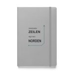 Silberfarbenes Hardcover-Notizbuch „Zwischen Zeilen liegt mein Norden“ mit schwarzer Schrift und petrolfarbener Navigationslinie, Vorderansicht.