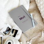 Silberfarbenes Notizbuch „Dein Mut schreibt weiter“ mit großer Schrift, schräg auf hellem Stoff fotografiert