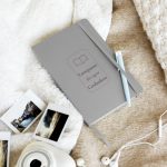 Silberfarbenes Lese-Notizbuch mit offenem Buchmotiv und Rahmen, schräg auf hellem Stoff fotografiert