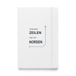 Weißes Hardcover-Notizbuch „Zwischen Zeilen liegt mein Norden“ mit schwarzer Schrift und petrolfarbener Navigationslinie, Vorderansicht.