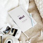 Weißes Notizbuch „Dein Mut schreibt weiter“ mit pflaumenfarbener Schrift, schräg auf hellem Stoff fotografiert